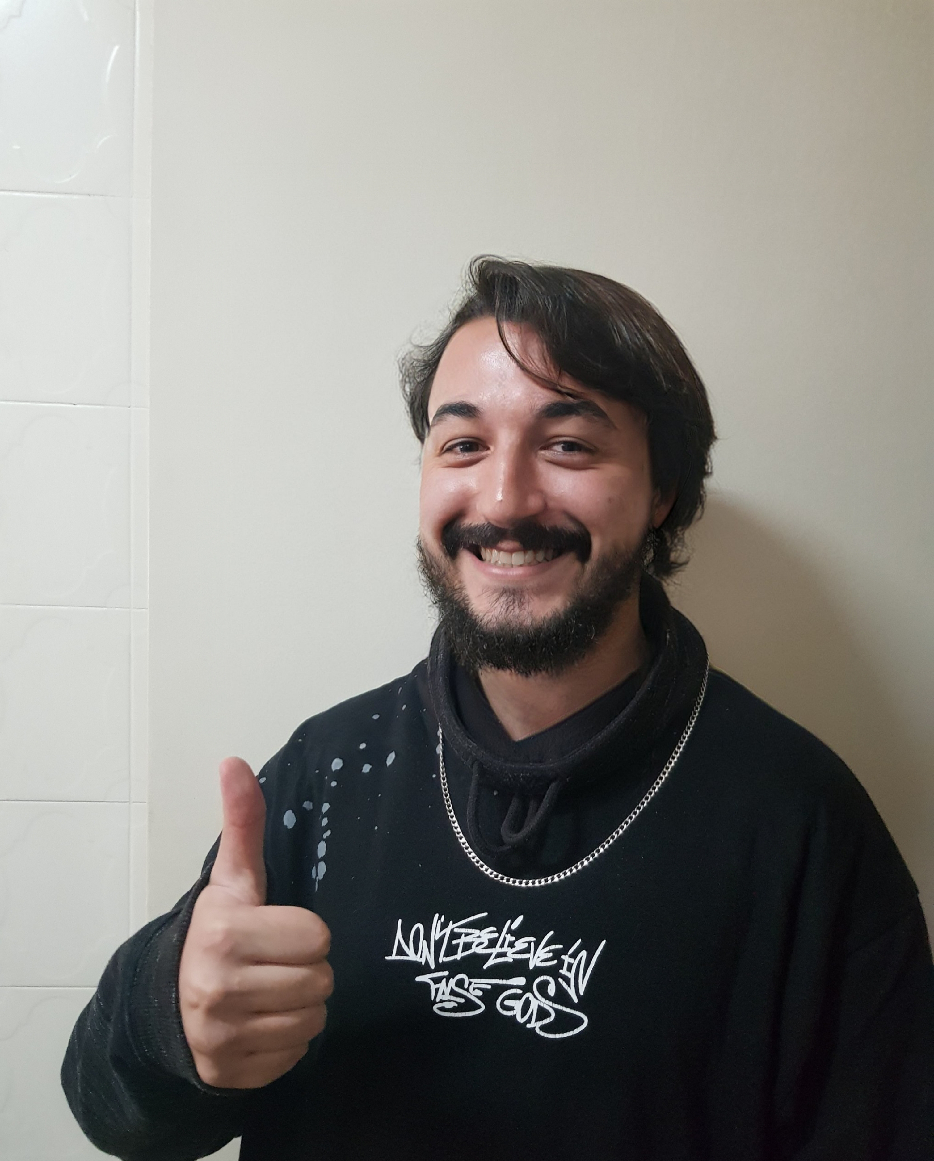 Foto de Tiago, ele está de casaco moletom preto escrito "Don't believe in false Gods", corrente. Está sorrindo e fazendo um joinha com mão direita